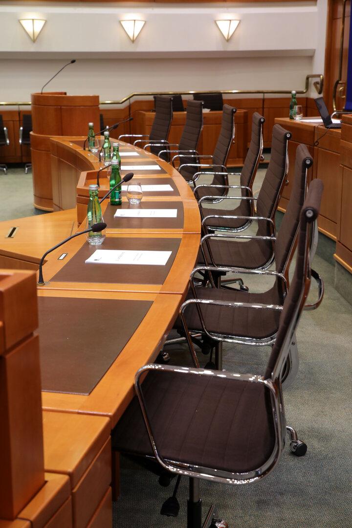 Leere Konferenzrunde mit Holz­tisch, Mikrofonen, Wasserflaschen und schwarzen Stühlen im Sitzungssaal.