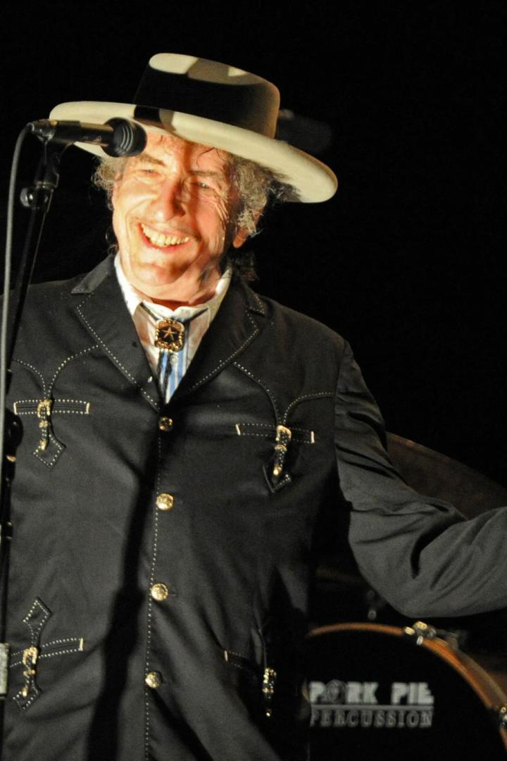 Bob Dylan lächelt auf einer Bühne vor einem Schlagzeug, trägt einen Hut und eine schwarze Jacke mit Bolo-Krawatte und hält ein Mikrofon in der Hand.