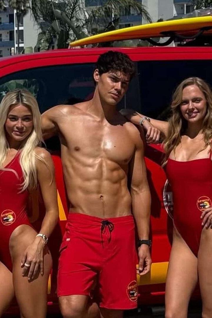 Fünf Rettungsschwimmer in roten Badeanzügen und Shorts posieren vor einem roten Lifeguard-Pick-up mit Surfbrettern auf dem Dach.