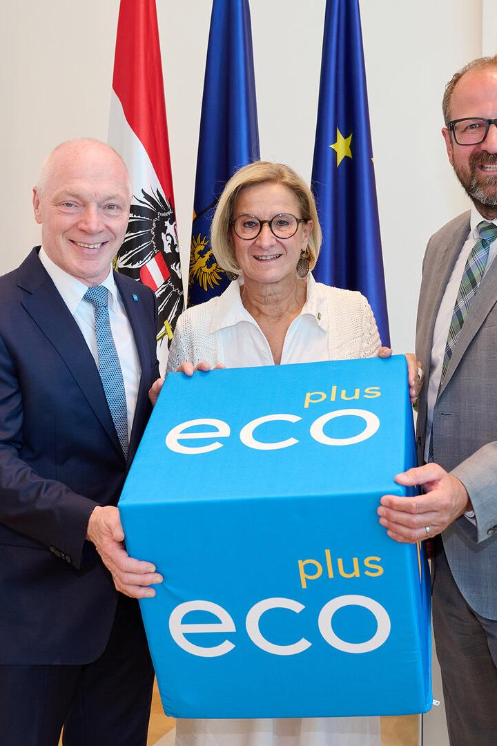 Drei Personen halten gemeinsam einen blauen „eco plus“-Würfel vor Fahnen in einem Büro.