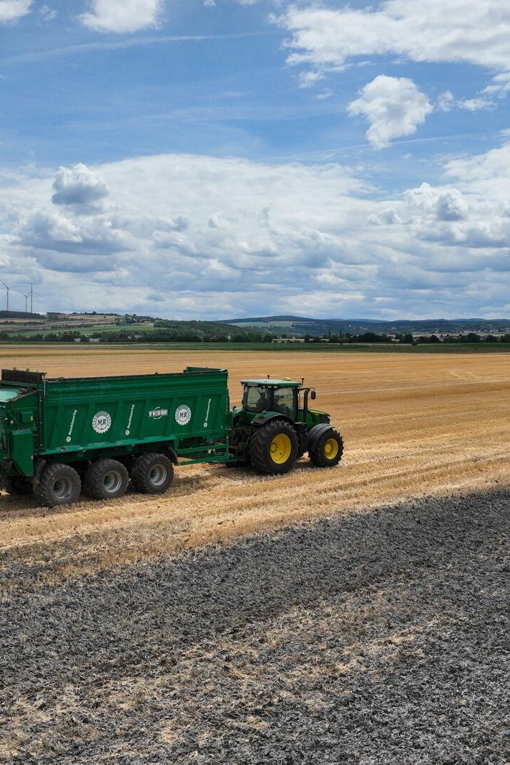 Traktor mit grünem Anhänger fährt über ein abgeerntetes Feld unter wolkigem Himmel.