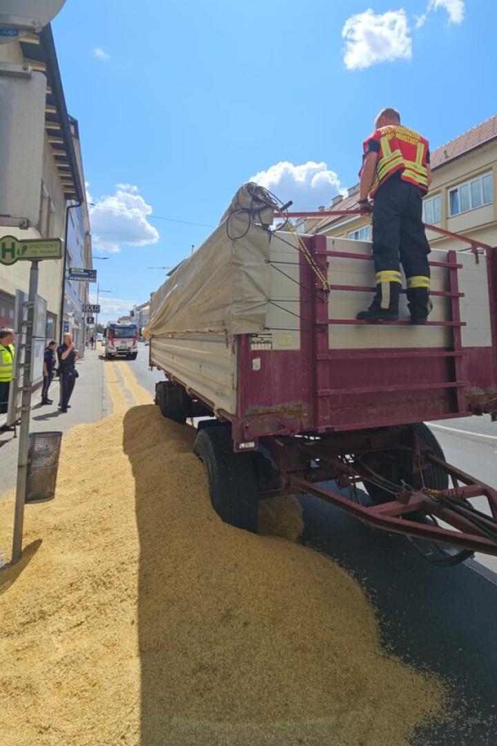 Ein Traktor mit Anhänger hat Maiskörner auf die Straße verloren, während ein Feuerwehrmann darauf steht.