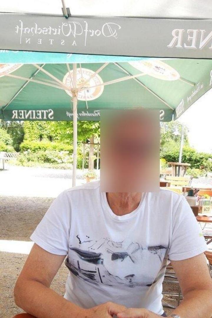 Eine Person sitzt im Freien an einem Tisch unter einem großen Sonnenschirm; das Gesicht ist unkenntlich gemacht.