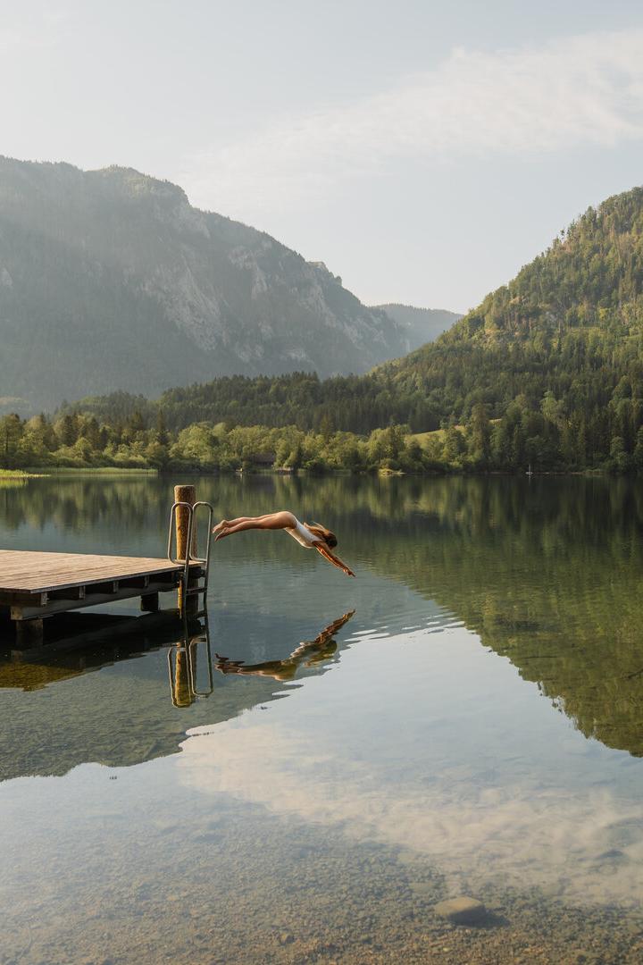 Eine Frau springt von einem Steg in einen stillen Bergsee, umgeben von bewaldeten Hängen und Spiegelungen im Wasser.