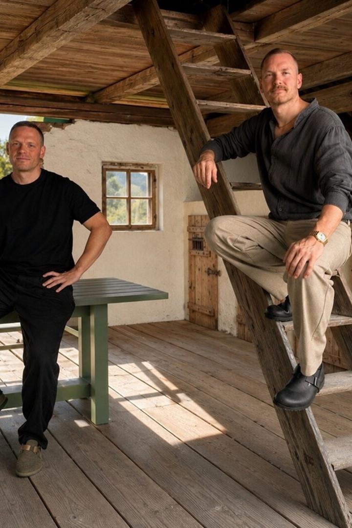 Zwei Männer posieren auf einer rustikalen Holzterrasse, einer sitzt am Tisch, der andere auf einer Leiter.