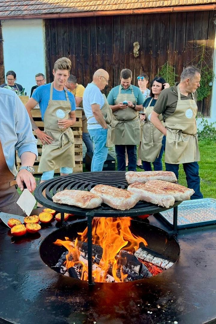 Mann grillt Fleisch und Gemüse an einer offenen Feuerstelle, während mehrere Personen in Schürzen zuschauen.