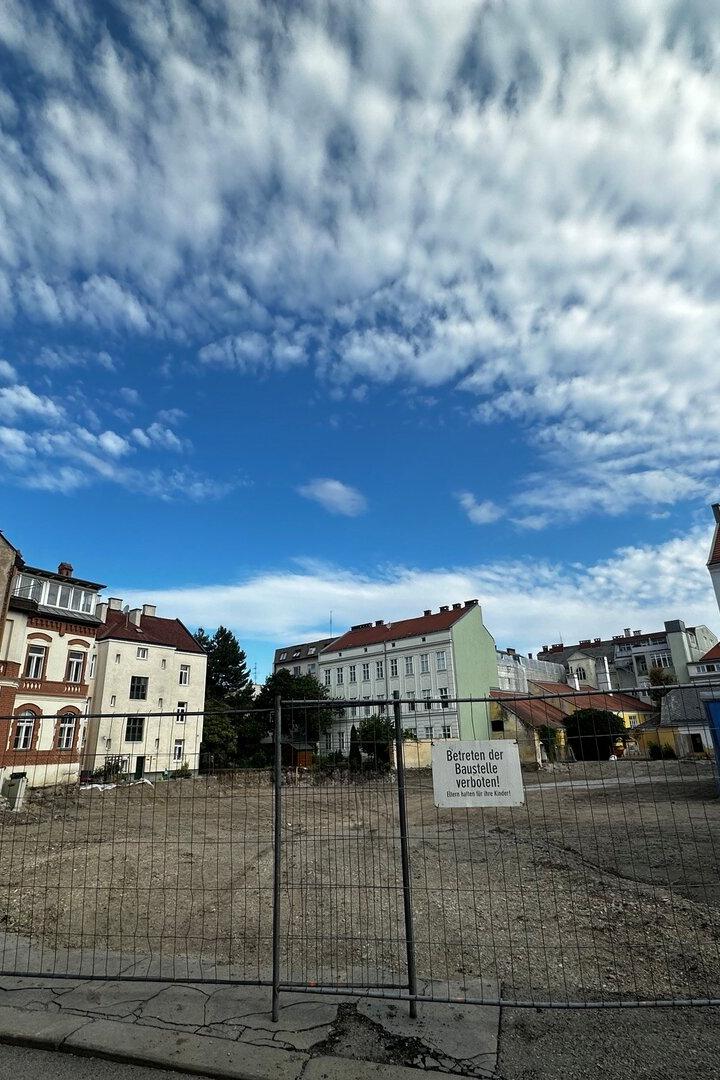 Umzäunte Baustelle zwischen Wohnhäusern unter dramatischem Wolkenhimmel.