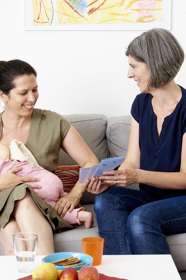 Eine Frau mit einem Säugling sitzt auf einem Sofa, daneben sitzt eine Familienbegleiterin, die ihr Informationsmaterial zeigt.