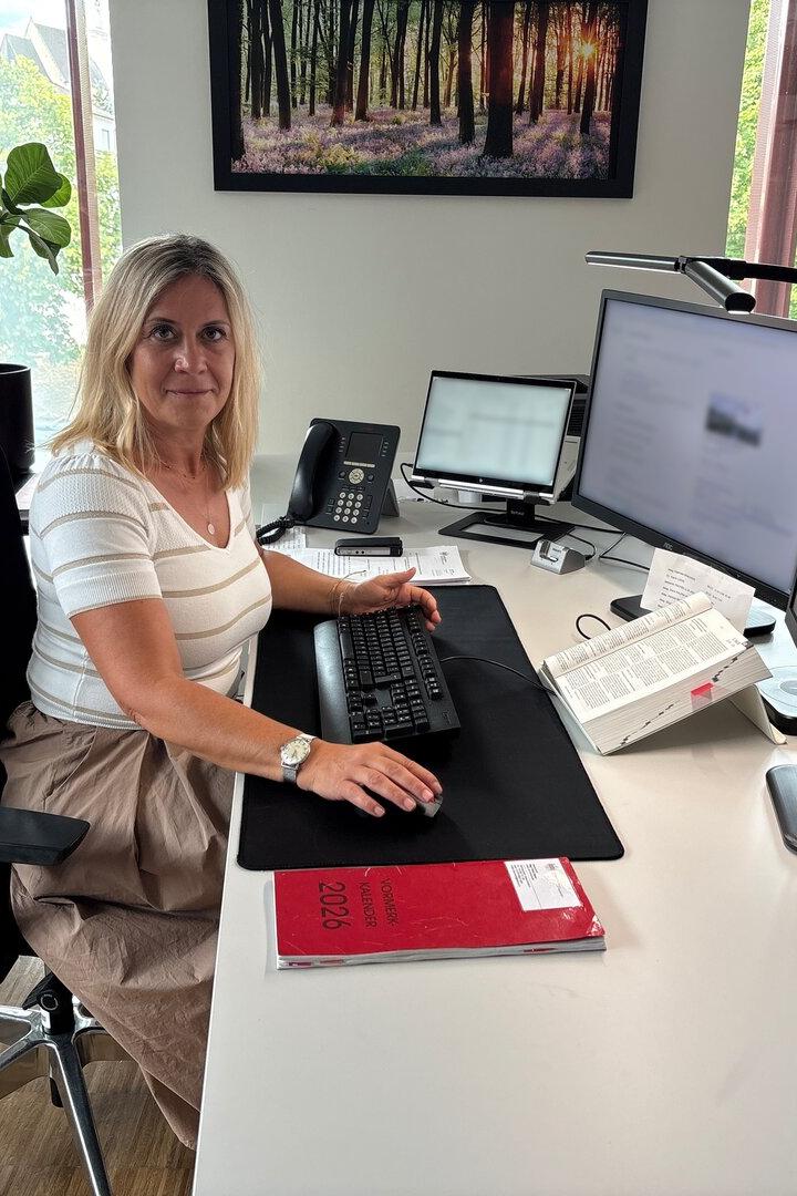 Frau sitzt an einem modernen Schreibtisch im Büro mit zwei Monitoren und Tastatur, umgeben von Pflanzen und Tageslicht.