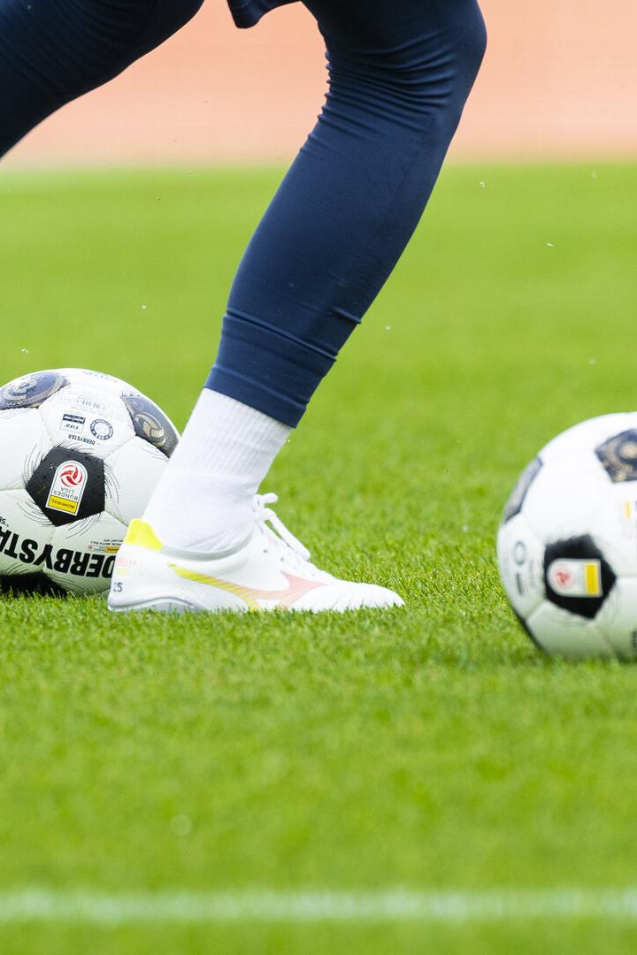 Fußballspiel mit zwei Bällen und den Beinen einer Spielerin auf dem Rasen.