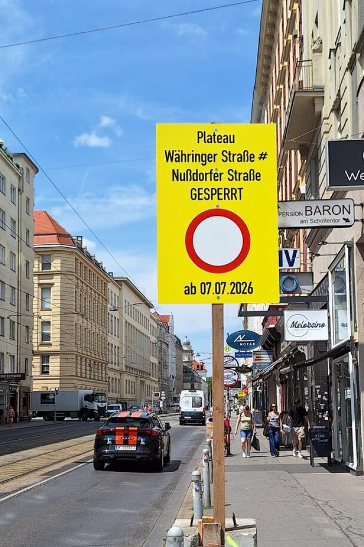 Gelbes Straßenschild kündigt die Sperrung der Währinger und Nußdorfer Straße ab 07.07.2026 an, an einer Wiener Straße mit Autos und Fußgängern.