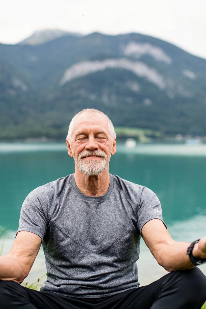 A senior man pensioner sitting by lake in nature, doing yoga exercise.