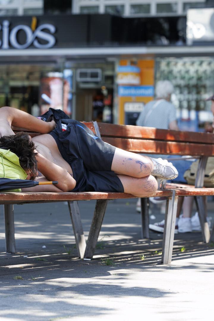 Eine Person schläft zusammengerollt auf einer Parkbank im Schatten.