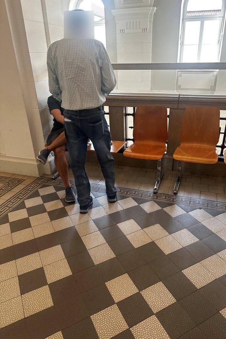 Zwei Männer stehen in einem hellen Flur neben einer Sitzreihe mit Holzstühlen.