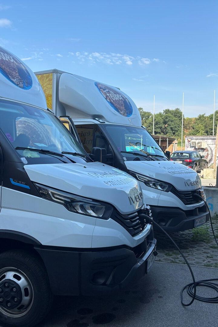 Zwei weiße Elektro-Lieferwagen der Bäckerei Felber stehen an einer blauen Wand und werden an Ladestationen aufgeladen.