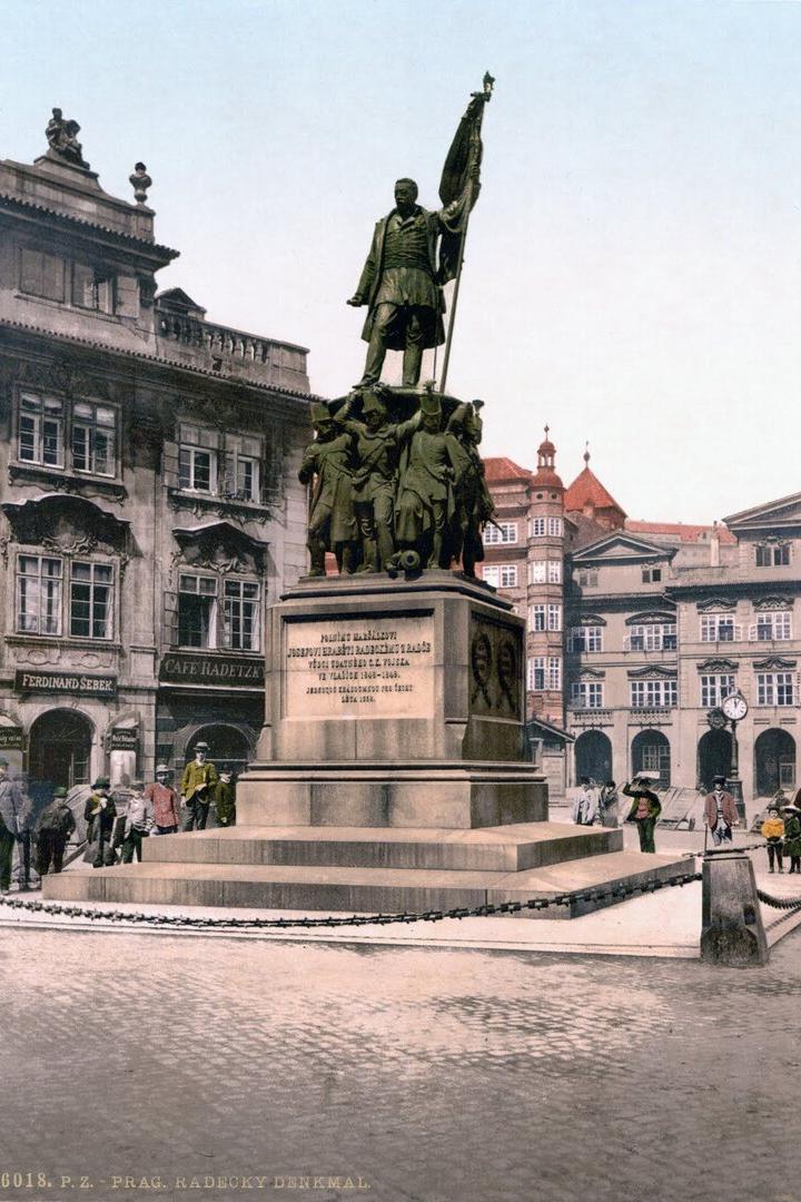 Denkmal eines Generals auf hohem Sockel in einem historischen Prager Platz, umgeben von alten Gebäuden und Passanten.