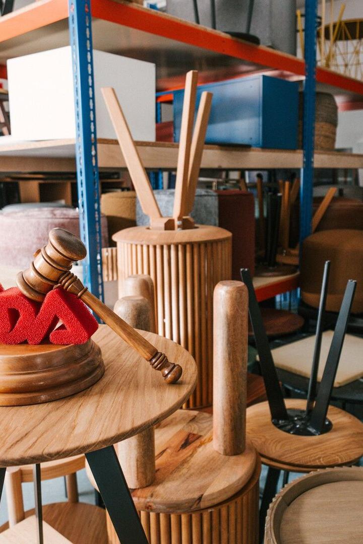 Ein Lager voller gestapelter Holzmöbel und Stühle, vorne ein Holzhammer auf einem Tisch mit roten ADA-Buchstaben.