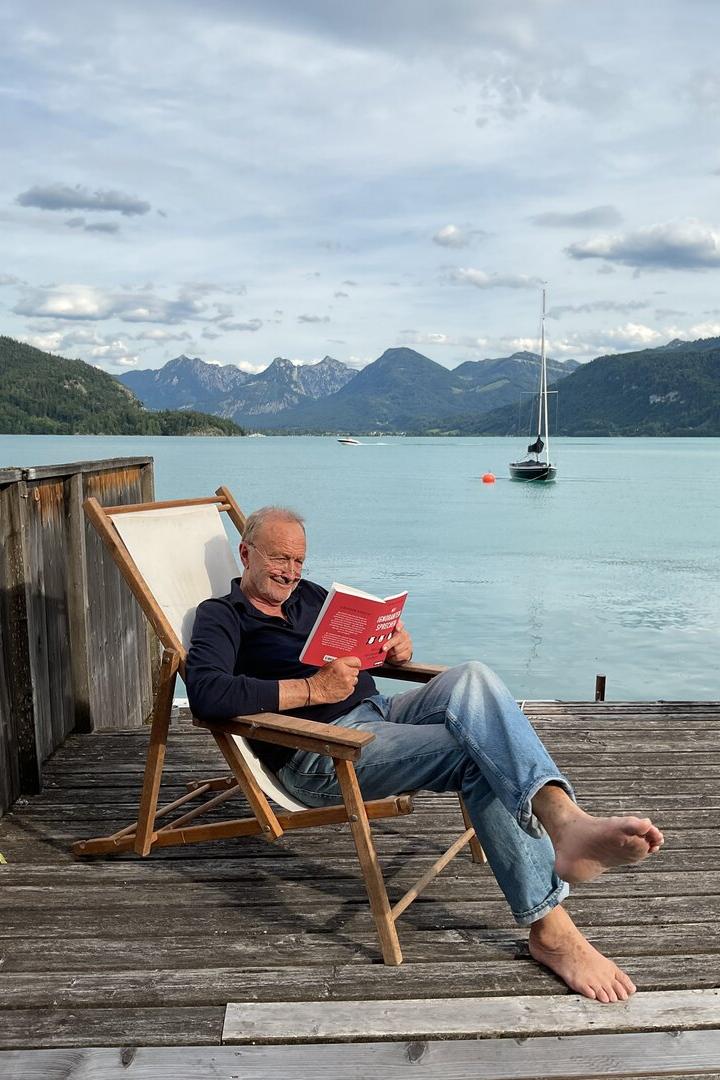 Ein Mann liest entspannt auf einer Holzterrasse am See, mit Segelboot und Bergen im Hintergrund.