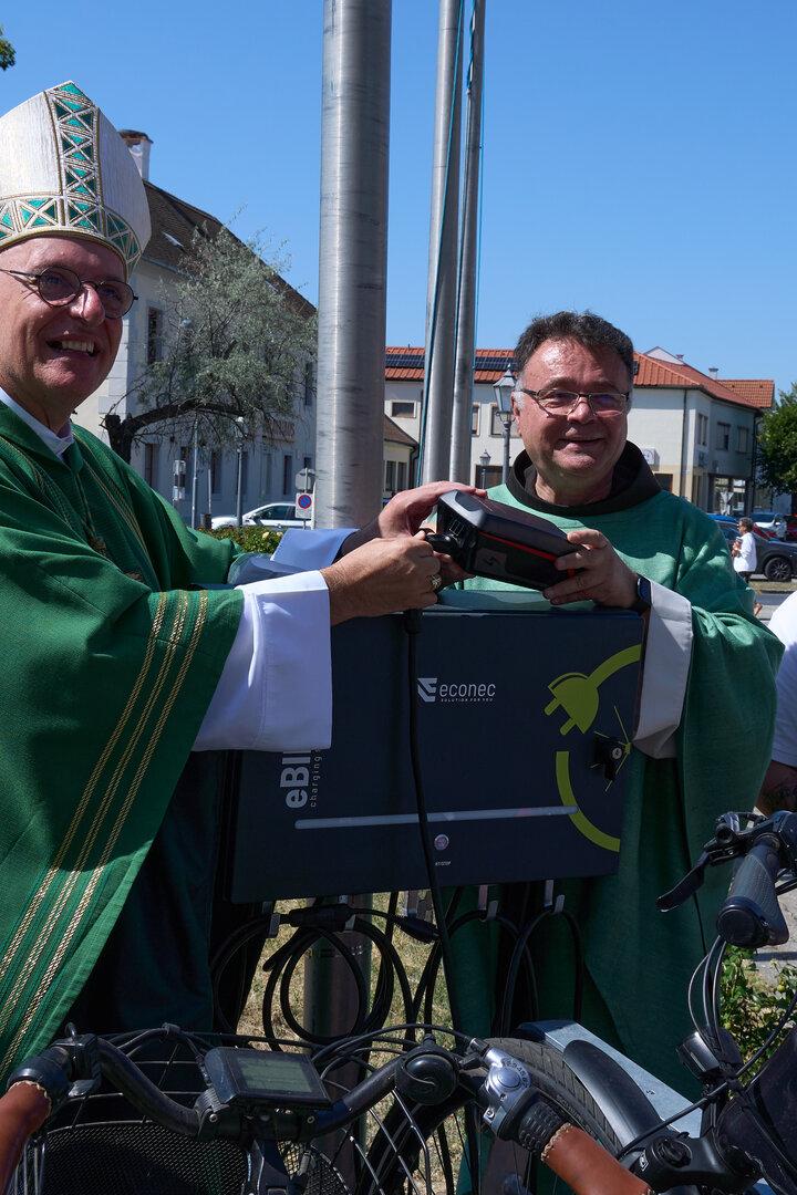 Ein Bischof segnet ein Fahrrad-Ladegerät, umgeben von Helfern und Fahrrädern im Freien.