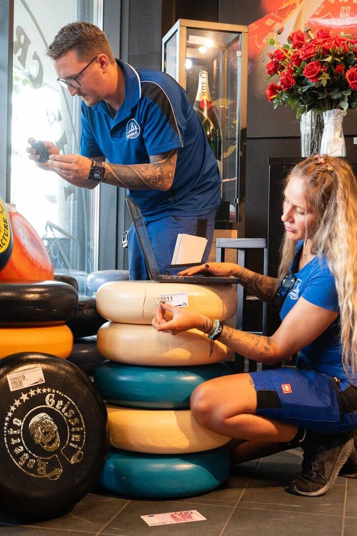 Zwei Personen in blauer Arbeitskleidung inventarisieren in einem Geschäft mehrere farbige Käselaibe von The Crazy Cheese Manufacture.