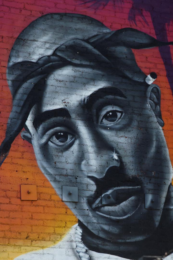 Graffiti-Porträt von Tupac Shakur auf einer Ziegelwand mit farbigem Hintergrund.