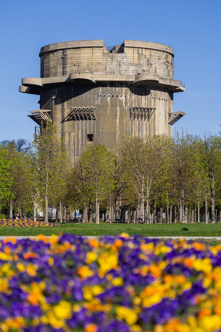 Der Wiener Flakturm im Augarten ragt hinter einer blühenden Blumenwiese und Bäumen in den blauen Himmel.