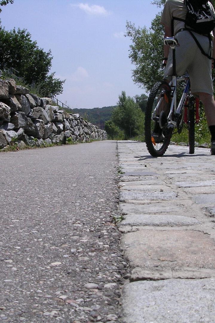 Ein Radfahrer mit Rucksack steht auf einem Weg aus Kopfsteinpflaster neben einer asphaltierten Strecke und einer Steinmauer.