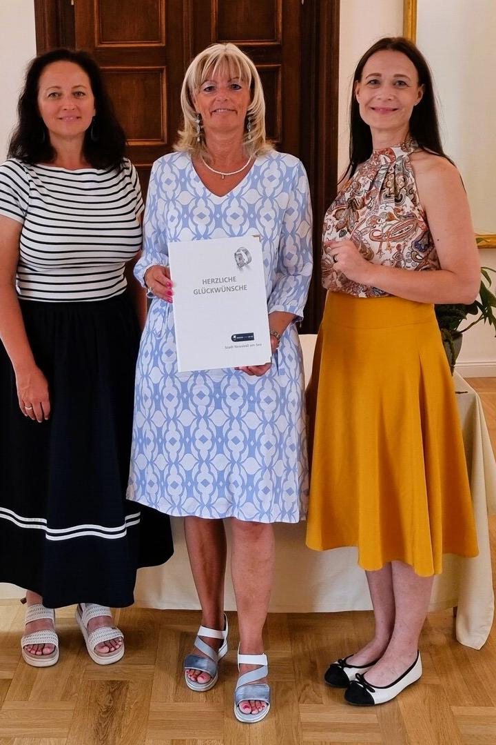 Drei Frauen (in der Mitte Stadtchefin Böhm) stehen nebeneinander, wobei die mittlere eine Urkunde der Stadt Neusiedl am See hält, flankiert von zwei weißen Herz-Dekorationen.
