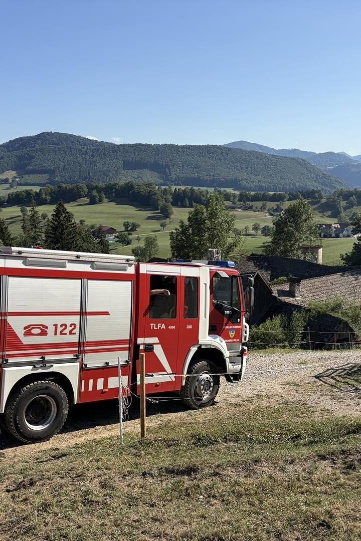 Ein rotes Feuerwehrfahrzeug steht auf einem Feldweg vor einer ländlichen Bergkulisse.