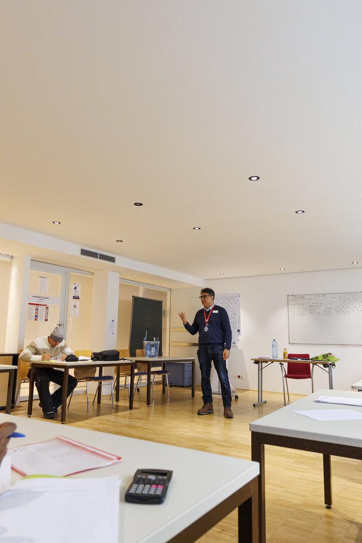 Mehrere Erwachsene sitzen in einem hellen Seminarraum an Tischen, während ein Dozent vorne spricht und an der Tafel steht.
