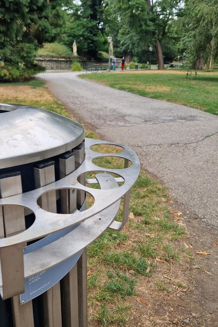 Ein Parkweg führt an einem großen Metall-Mülleimer vorbei, im Hintergrund stehen Bäume und vereinzelte Spaziergänger.