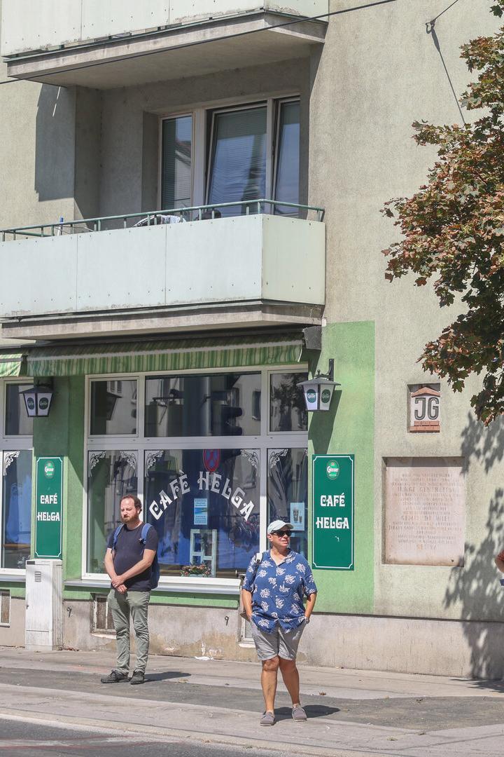 Straßenszene vor dem Café Helga mit mehreren wartenden Passanten, Balkon, Baum und Bushaltestelle.