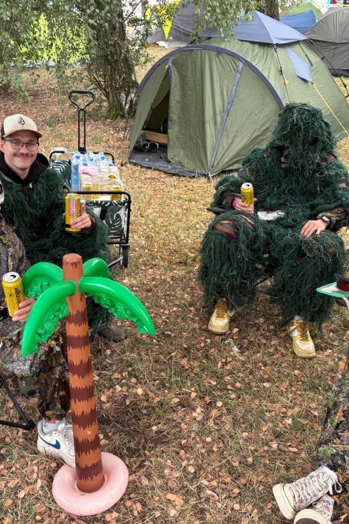 Vier als Waldmenschen verkleidete Frequency-Besucher sitzen mit Bierdosen vor Zelten, neben einer Plastikpalme auf dem Campingplatz.