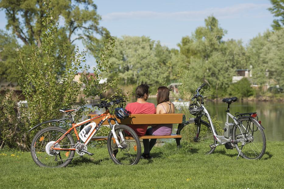 Ein Paar sitzt auf einer Bank am See, flankiert von zwei Fahrrädern.