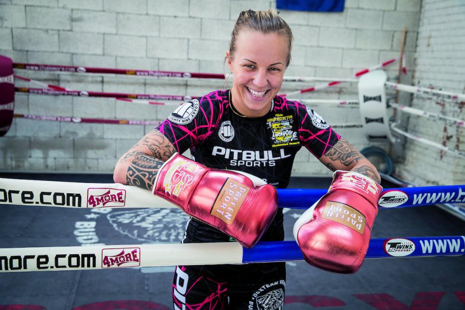 Eine tätowierte, lächelnde Frau mit Boxhandschuhen lehnt im Ring.