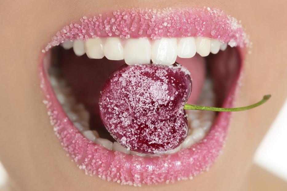 Eine Kirsche, bedeckt mit Zucker, wird von einem Mund mit ebenfalls gezuckerten Lippen gehalten.