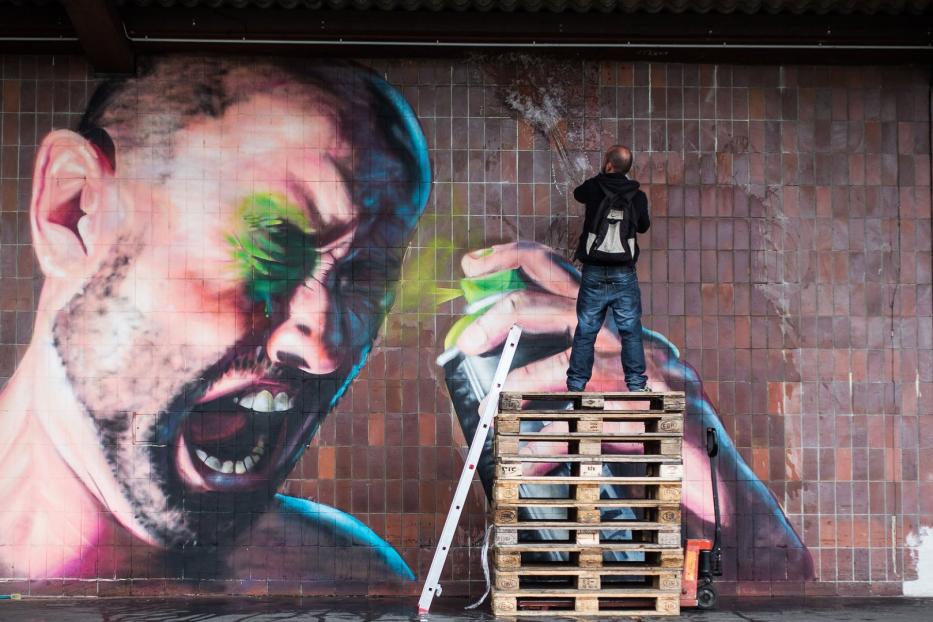 Ein Mann sprüht ein Graffiti auf eine Ziegelwand, das ein schreiendes Gesicht zeigt.