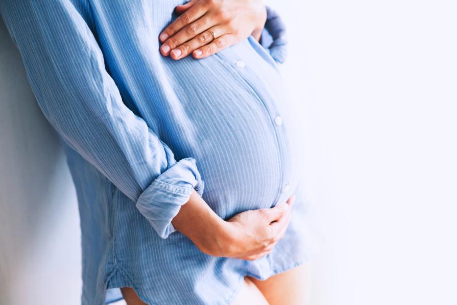 Eine schwangere Frau in einem hellblauen Hemd hält ihren Bauch.