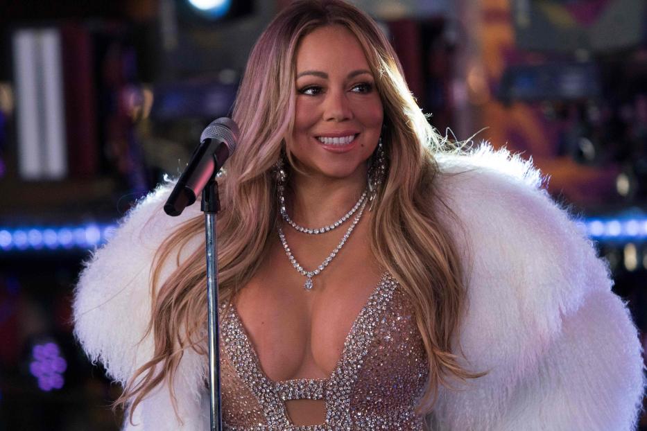 Mariah Carey steht lächelnd mit Glitzerkleid und weißem Fellmantel vor einem Mikrofonständer.