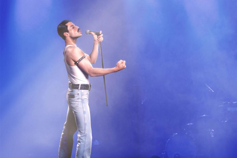 Ein Mann im Stil von Freddie Mercury singt mit einem Mikrofon vor blauem Hintergrund.