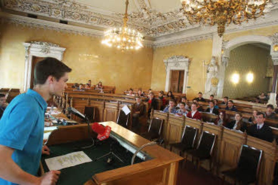 Ein junger Mann spricht vor einem Publikum in einem prunkvollen Saal.