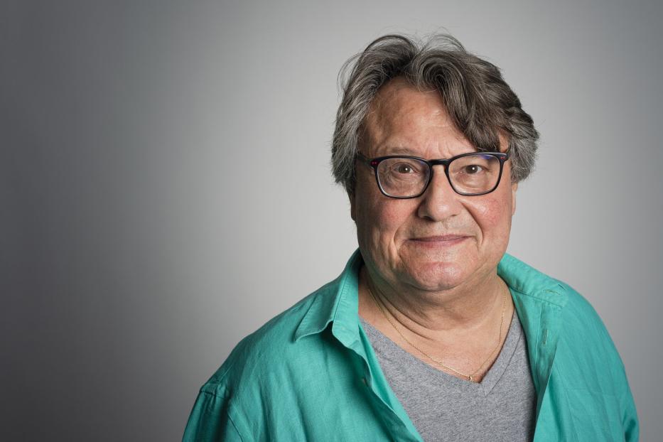 Ein Porträt eines Mannes mit Brille und einem grünen Hemd vor einem grauen Hintergrund.