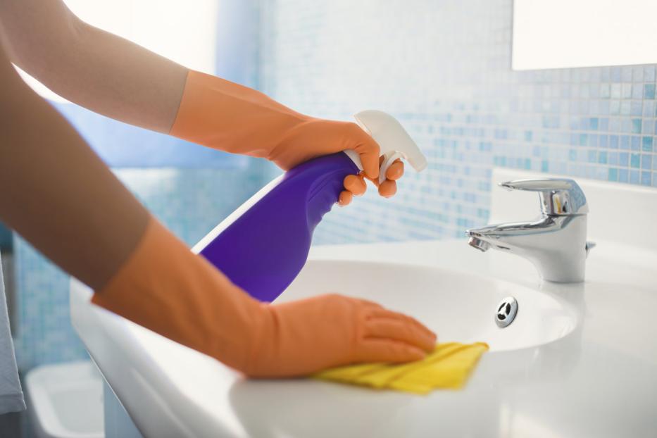 Eine Person mit orangefarbenen Handschuhen putzt ein Waschbecken mit einem gelben Tuch und einem Reinigungsspray.