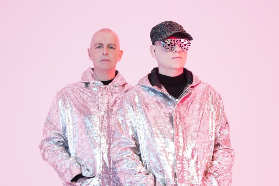 Die Pet Shop Boys in glänzenden Jacken vor einem rosa Hintergrund.
