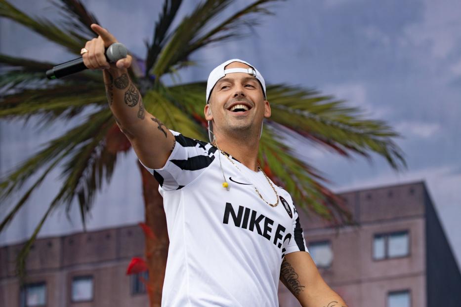 Kontra K tritt in einem Nike-Trikot mit Tätowierungen und einer Kappe auf einer Bühne auf.