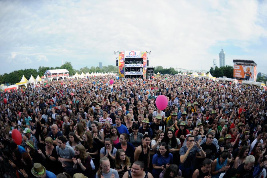 Eine große Menschenmenge jubelt vor einer Bühne von Hitradio Ö3 mit dem Donauturm im Hintergrund.