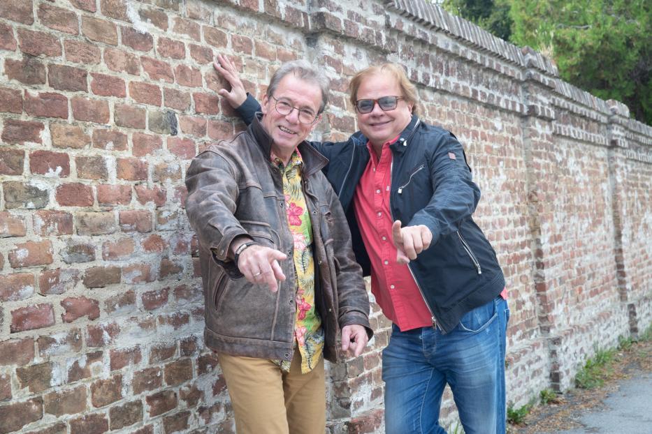 Zwei Männer posieren lächelnd vor einer Backsteinmauer.