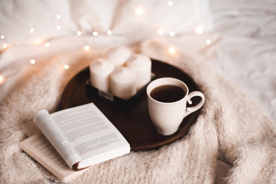 Eine Tasse Tee, ein Buch und Kerzen auf einem Tablett im Bett.