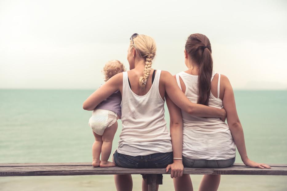 Zwei Frauen und ein Kleinkind sitzen auf einer Bank mit Blick auf das Meer.