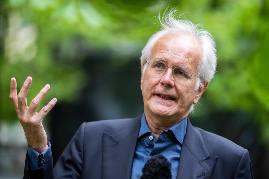 Ein Mann mit weißem Haar und Brille gestikuliert vor einem grünen Hintergrund.
